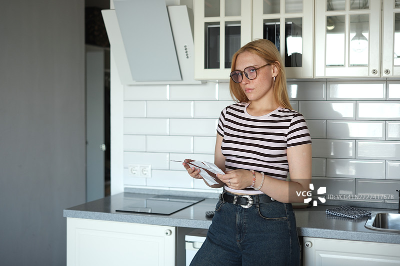 忧虑的Z世代女孩在厨房里看着账单，感到压力，年轻女性在为金钱而苦恼，计算每月开支。生活成本上升，学生贷款债务。图片素材