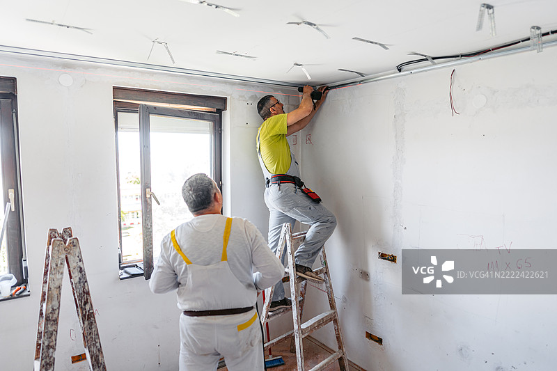 两名成熟工人在空置的正在装修的公寓里用电钻在天花板上安装石膏板型材图片素材