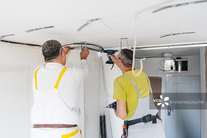 两名成熟工人在空置的正在装修的公寓里用电钻安装石膏板天花板。图片素材