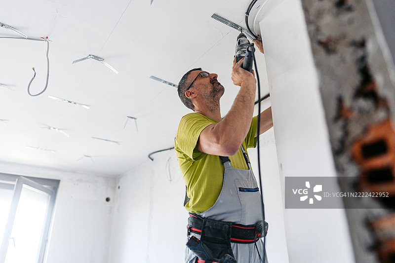 成熟工人在空置的翻新公寓的天花板上用电钻安装干墙间隔器图片素材