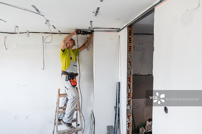 成熟工人在空置的正在装修的公寓里用电钻安装石膏板型材于天花板上图片素材