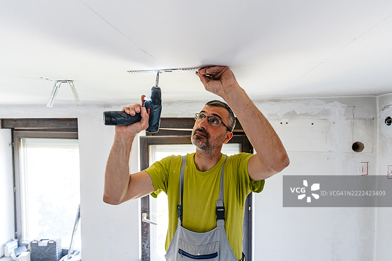 成熟工人在空置的翻新公寓天花板上用电钻安装干墙间隔器图片素材