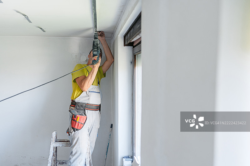 成熟工人在空置的公寓天花板上用电钻安装石膏板型材，公寓正在装修中。图片素材