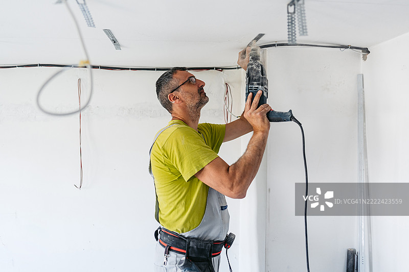 成熟工人在空置的翻新公寓里用电钻在天花板上安装干墙间隔。图片素材