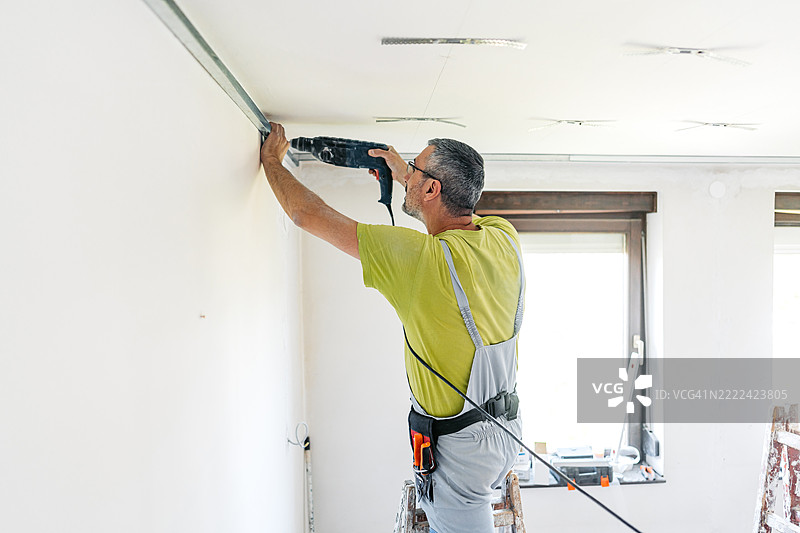 成熟工人在空置的公寓天花板上用电钻安装石膏板型材，公寓正在装修中。图片素材