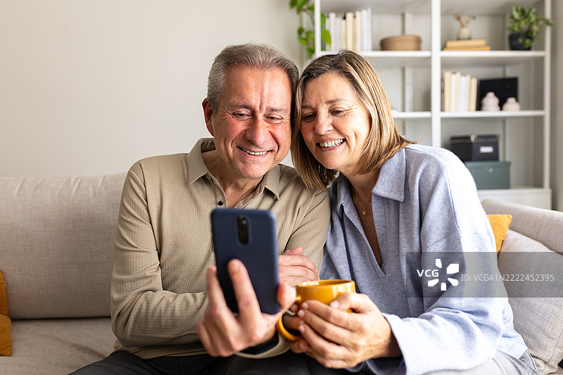 快乐的老年夫妇在沙发上使用智能手机并喝咖啡图片素材