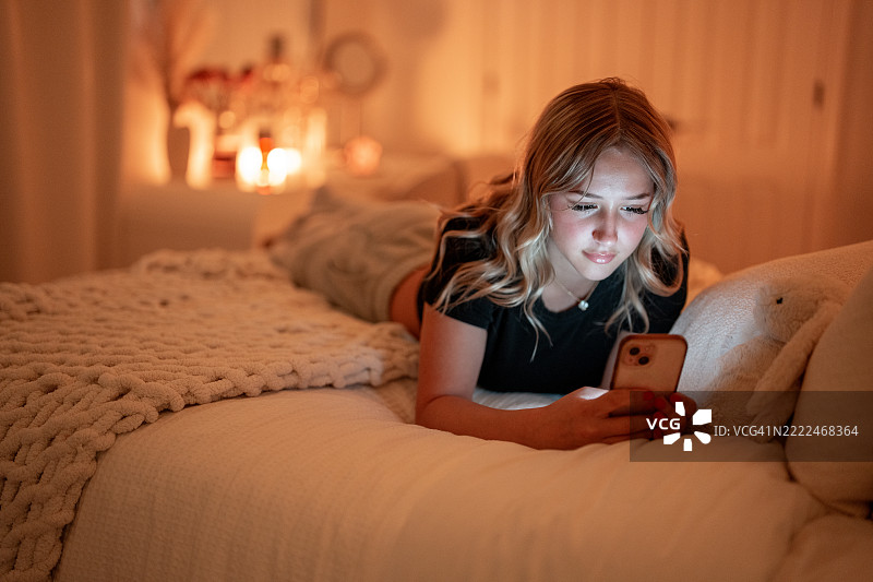年轻女性在温馨昏暗的房间里放松，使用智能手机躺在床上图片素材