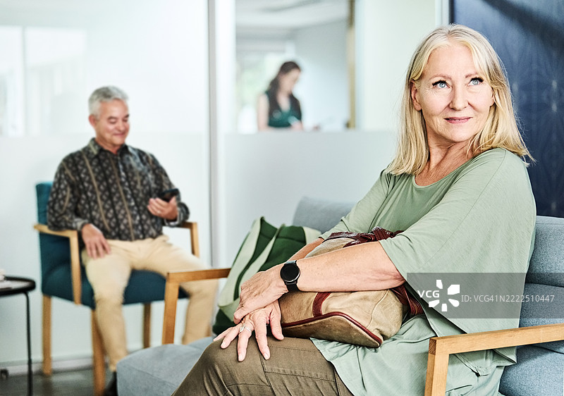 一位老年女性坐在诊所的候诊室，背景中有一名男性正在使用手机。图片素材