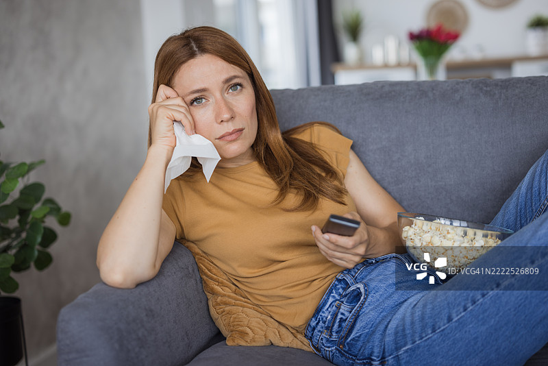 伤心的年轻女性在看电视，擦拭眼泪用纸巾。图片素材