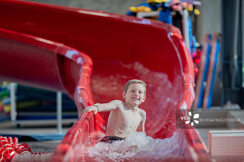 年轻男孩在室内游泳池的红色滑水道上滑下。图片素材