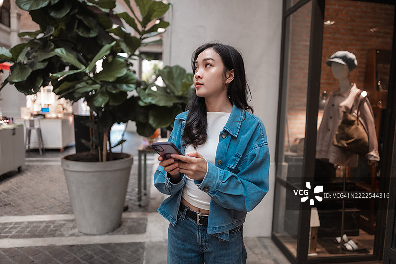 亚洲女性在繁华的购物中心欣赏商店橱窗时，边浏览智能手机，享受城市购物的一天图片素材