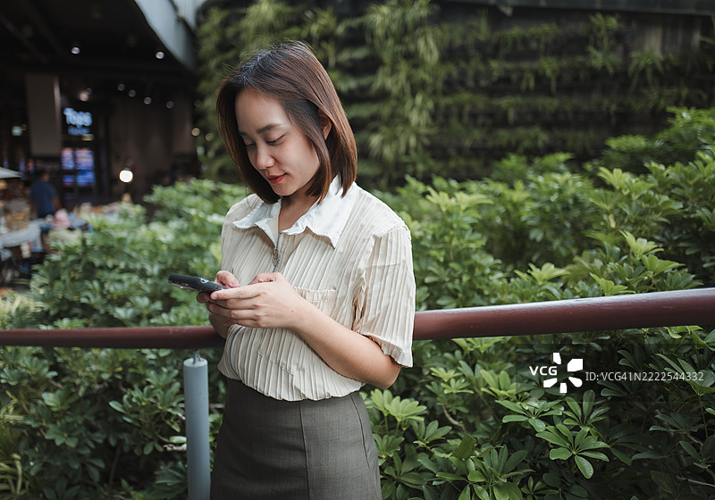 年轻的亚洲女商人在享受绿色城市空间时，使用智能手机发短信，周围环绕着植物和树木。图片素材