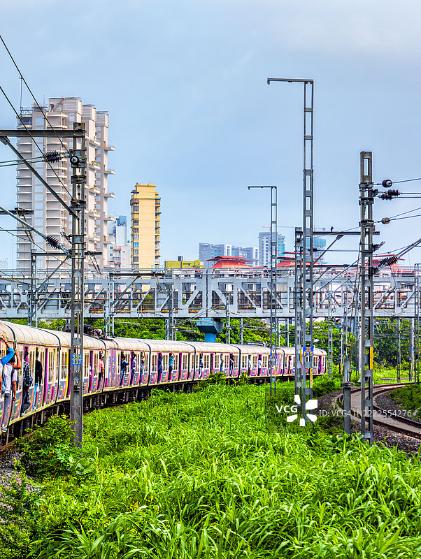 一列拥挤的孟买本地列车在高楼大厦和电气铁路基础设施的背景下,穿行于生机勃勃的绿色走廊。照片捕捉到列车沿着轨道弯曲,乘客们从车门外探出身子。图片素材