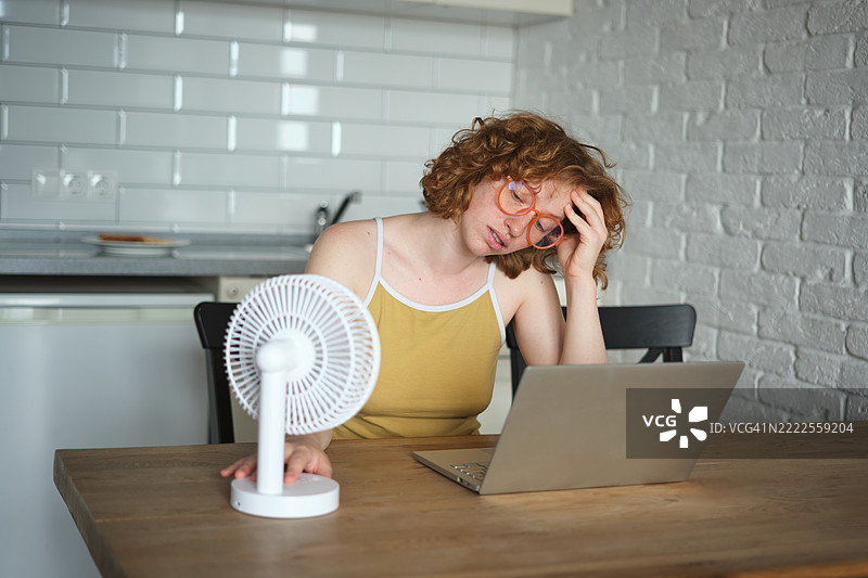 在热浪中工作。女性自由职业者在夏季炎热中感到不适，坐在厨房桌子旁，居家在线工作，使用电风扇。图片素材