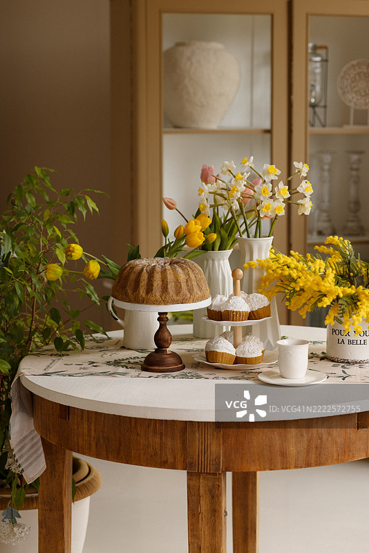 温暖经典房间的乡村圆桌上摆放着春天的花朵和甜点，旁边有木制橱柜。图片素材