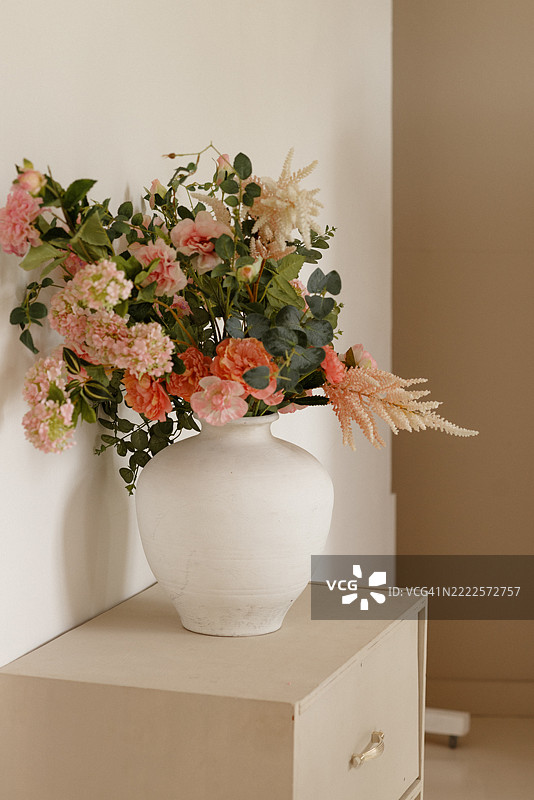 白色陶瓷花瓶中盛放着粉色和桃色花卉的花艺作品，摆放在米色复古橱柜上。图片素材