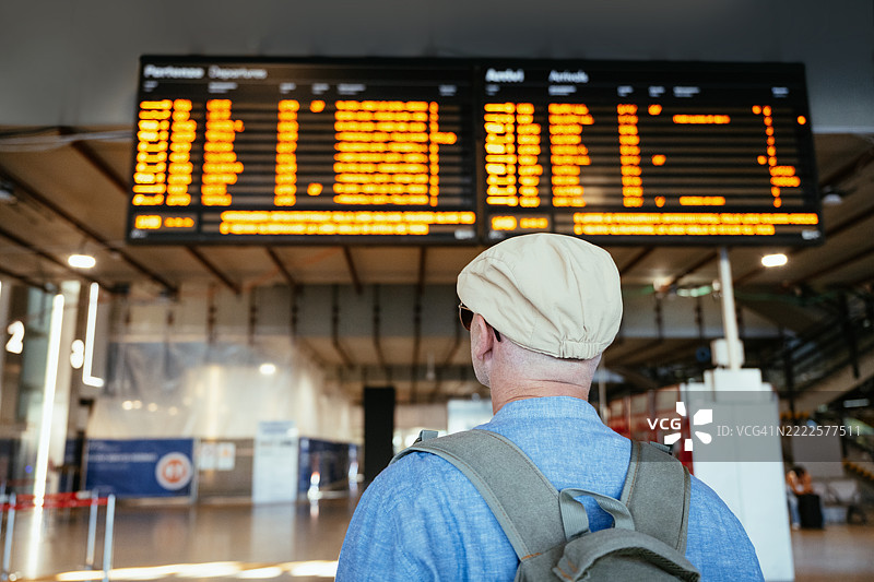 微笑的男性旅行者在火车站，手持智能手机，背着背包，穿着休闲服，背景有电子时刻表。图片素材