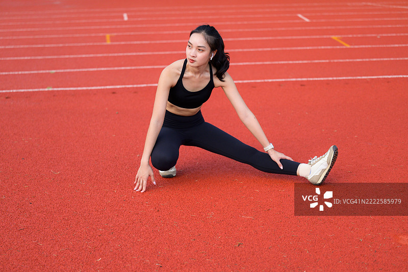 年轻的亚洲女性在鲜艳的红色跑道上拉伸腿部，展现出她在健身训练中的专注和奉献。图片素材