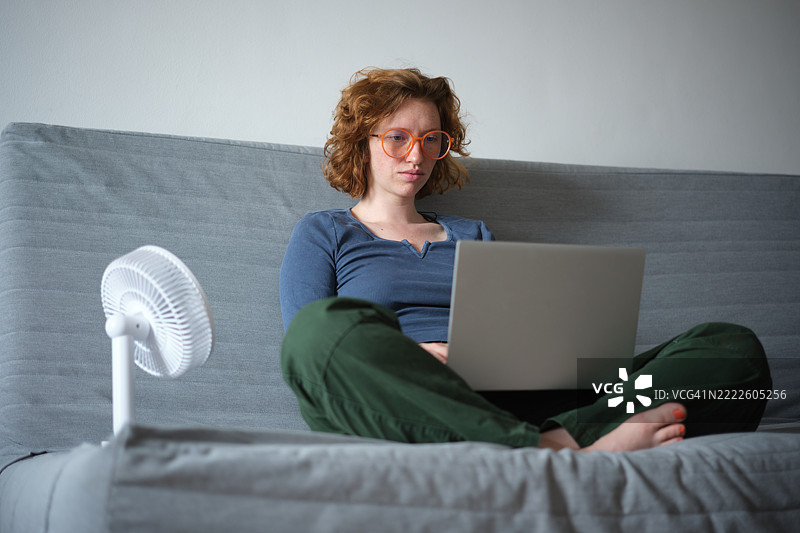 疲惫的女性自由职业者坐在沙发上使用笔记本电脑，家中使用电风扇，在热浪中远程工作，忍受夏季的炎热。保持家庭办公室在夏季炎热中凉爽。图片素材