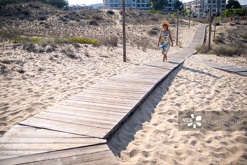 一位穿着色彩斑斓裙子和宽边草帽的美丽成熟的女性在海滩小径上漫步。图片素材