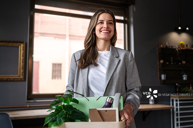 职业女性自信微笑着手持纸箱，离开公司办公空间，经历工作转变。图片素材