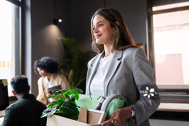 年轻女商人微笑自信地提着装有个人物品的箱子，离开办公室，辞去了工作。图片素材