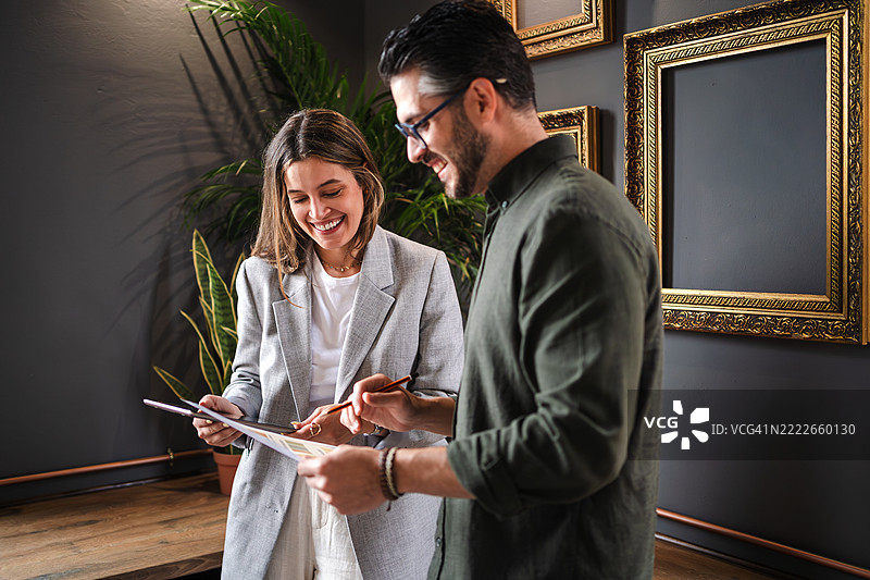 两位年轻的艺术画廊老板微笑着讨论商业策略，手中拿着平板和文件，周围是空的金色画框。图片素材