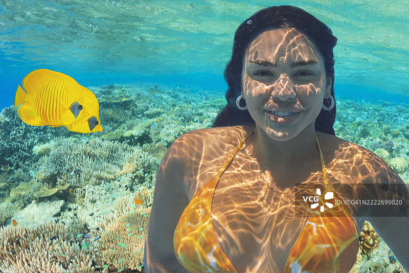 快乐的女性露出灿烂的笑容，和一对黄色鱼在一起，水下照片中有两只面具蝴蝶鱼和珊瑚海洋生物。图片素材