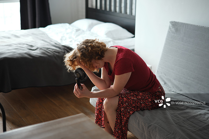 夏季热浪。年轻女性在家中因炎热而感到不适，使用便携式电动迷你风扇，享受清新空气，在热浪中降温。热潮概念图片素材