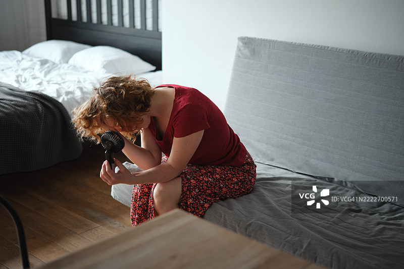 夏日炎热。卷发红发的女人在家中炎热的日子里坐在电风扇前。图片素材