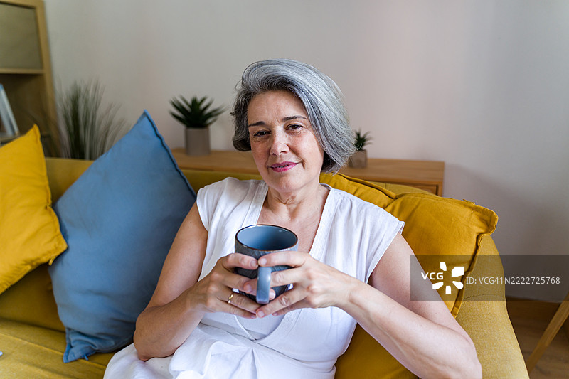 一位年长女性坐在沙发上放松，手里拿着一个杯子，面带微笑看向镜头。图片素材
