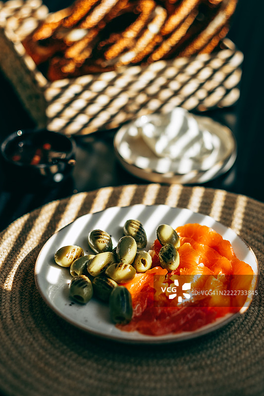 盘子里放着绿色橄榄和烟熏三文鱼片，置于编织的餐垫上。阳光与阴影交错。背景中有一碗奶油奶酪和一个面包篮。图片素材