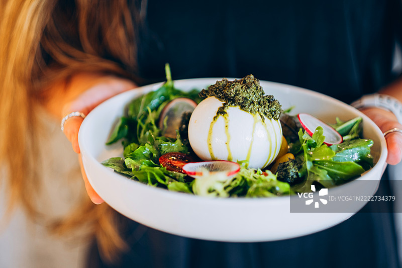 女人的手拿着白色盘子，上面放着新鲜的绿色沙拉、布拉塔奶酪、香蒜酱和切片的萝卜。女人有着长发，佩戴着休闲首饰，背景柔和模糊。图片素材
