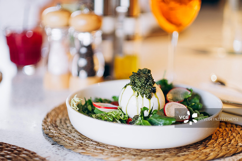 白色盘子里盛着新鲜的绿色沙拉、萝卜片、樱桃番茄和一整块撒上绿色香蒜酱的布拉塔奶酪。明亮的环境，自然光，编织的餐垫，休闲用餐。图片素材