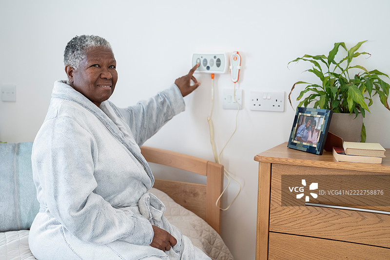 微笑的老年女性穿着浴袍坐在床上，按下墙上的紧急按钮图片素材
