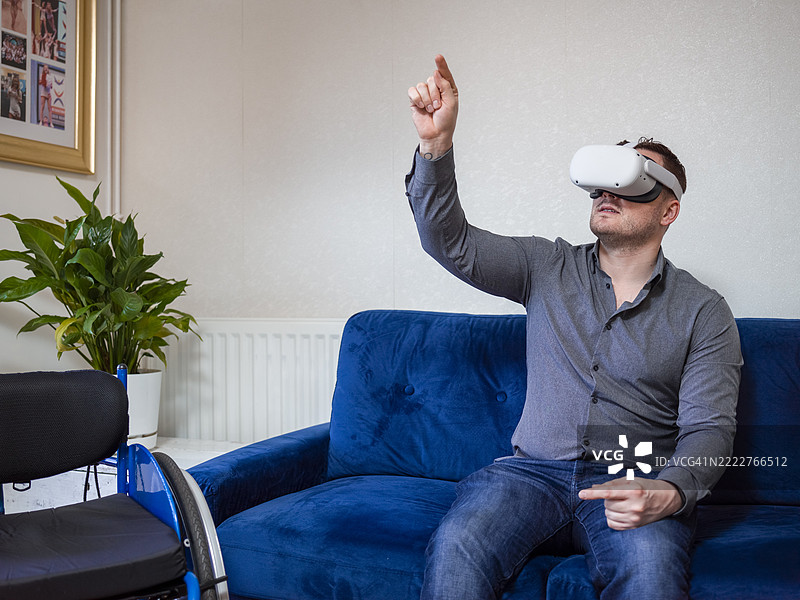 戴着虚拟现实眼镜的男子坐在沙发上，旁边有一把轮椅。图片素材