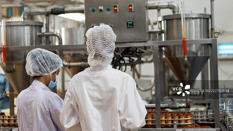 穿着发网和无菌制服的食品科学家和质量控制官员正在检查罐装鱼的生产线，以确保产品质量和安全。图片素材