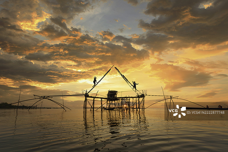 日出时，两名男子在泰国帕特塔隆的传统渔网架上攀爬的剪影图片素材