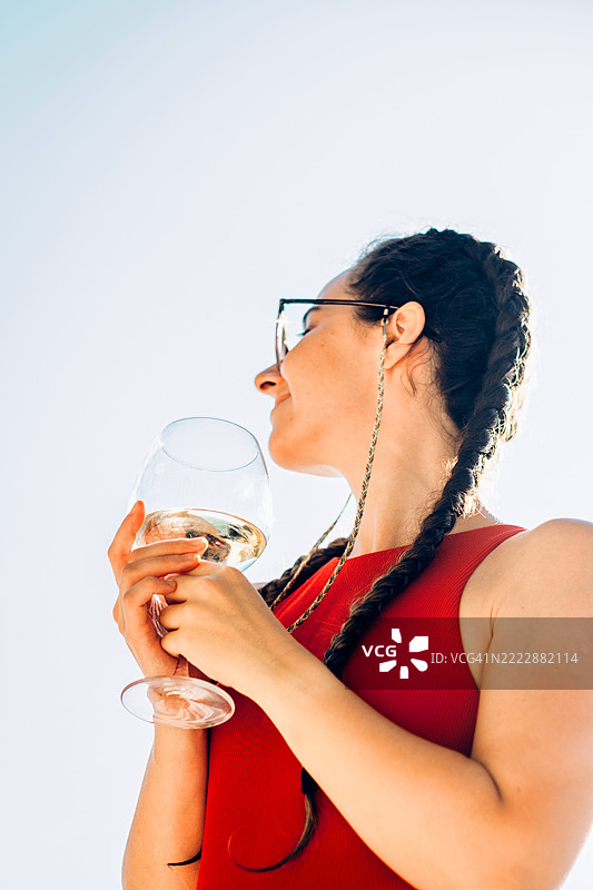 年轻美丽的女子扎着辫子，手持一杯白葡萄酒，朝一旁望去，沐浴在阳光下。夏季放松与户外饮品的概念。图片素材