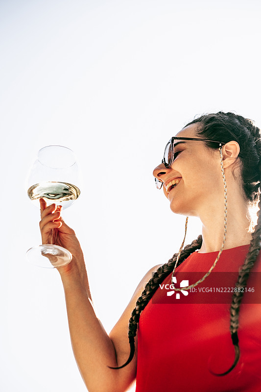 微笑的年轻女性扎着辫子，手中举着一杯白葡萄酒，背景是晴朗的天空。欢乐、庆祝和夏季饮品的概念。图片素材