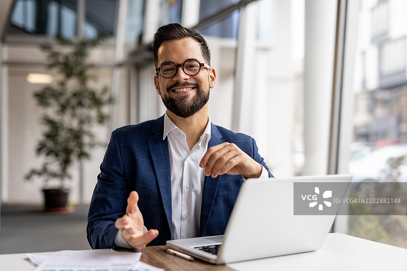 微笑的商人在桌子前坐着，手势表达，旁边有一台笔记本电脑。图片素材