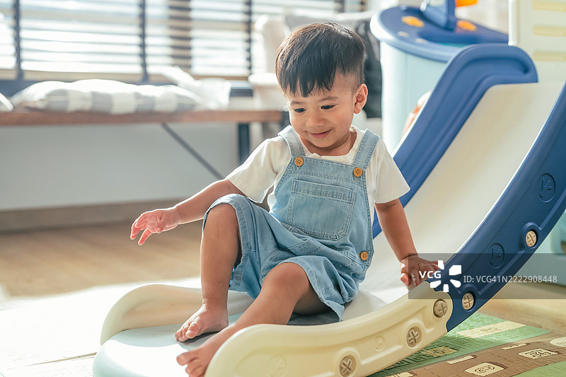 亚洲幼儿男孩在家中的游乐屋隧道和滑梯上爬行，快乐地玩耍，享受与塑料玩具屋的乐趣，展现出健康的生活方式。图片素材