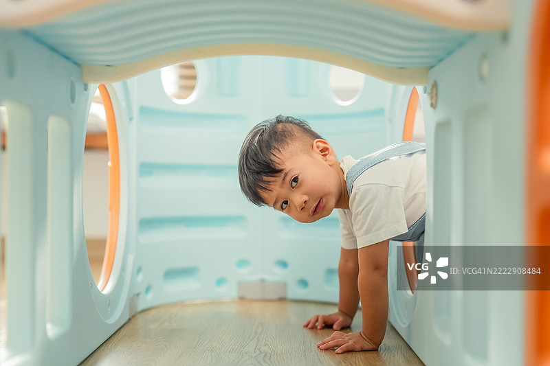 亚洲幼儿男孩在家中的游乐屋隧道和滑梯上爬行，快乐地玩耍，享受与塑料玩具屋的乐趣，展现出健康的生活方式。图片素材