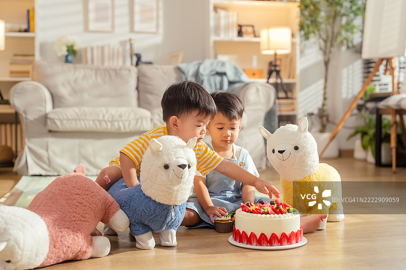 两个幼儿男孩快乐地坐在地板上一起庆祝生日。一个孩子正在吹灭五彩缤纷的生日蛋糕上的蜡烛，而另一个孩子则兴奋地用手指戳进奶油中，满脸开心。周围是气球和蛋糕屑。图片素材