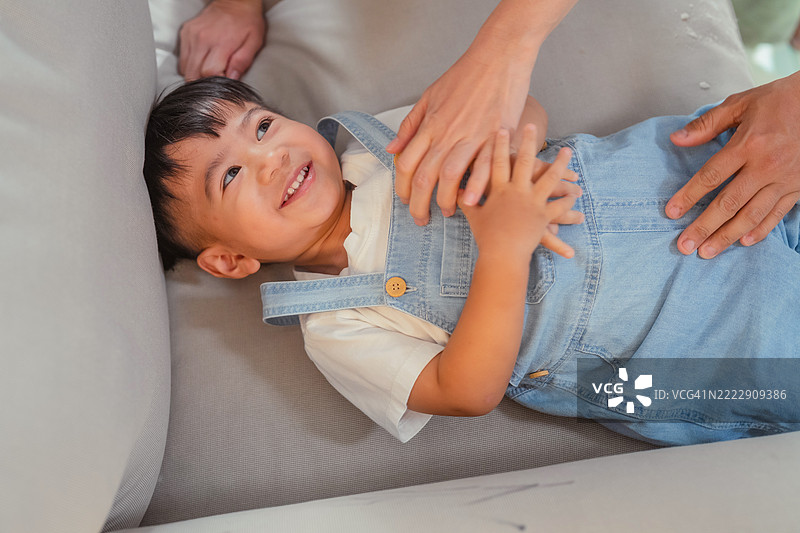 温馨的家中，两个小男孩在沙发上顽皮地爬上爬下，和爸爸嬉戏玩耍，快乐的笑声充满了整个房间，妈妈也加入其中，轻轻挠痒和拥抱儿子，营造出温暖而自然的团聚场景。图片素材