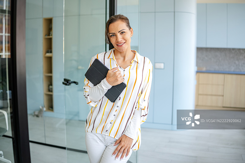 一位自信的女性在现代厨房中微笑着手持平板，阳光明媚。图片素材