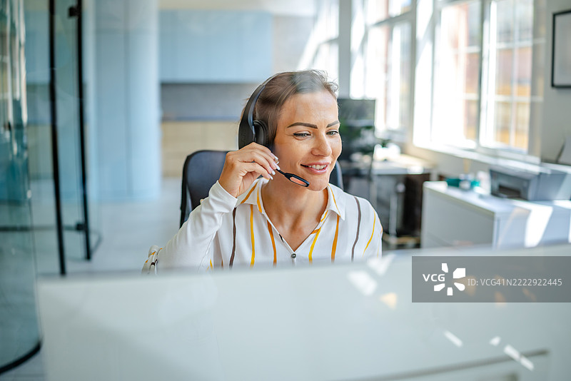 在现代办公室环境中，一位女性佩戴耳机，面带微笑，坐在电脑屏幕前工作。图片素材