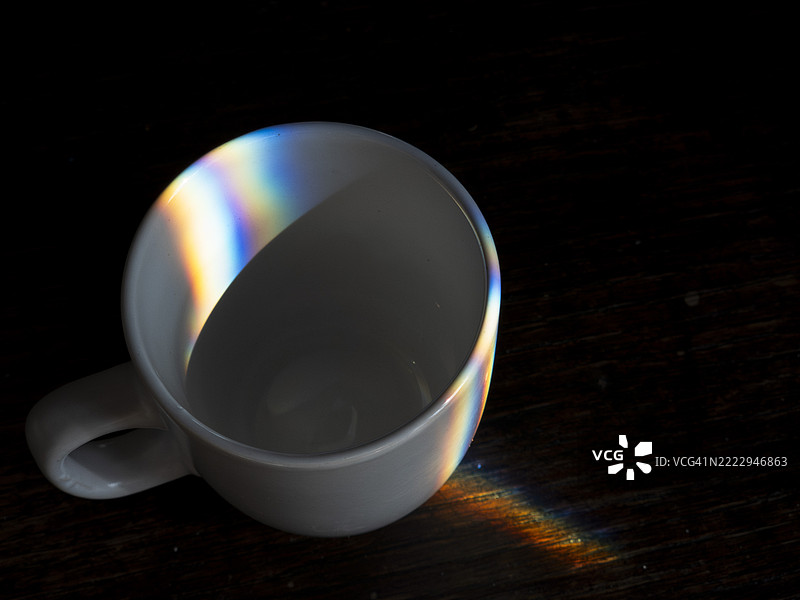 光线透过一个空白色杯子在深色木质表面上折射，营造出一种微妙的彩虹效果，增添了温馨的氛围。图片素材
