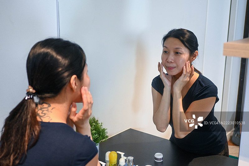 美丽的亚洲女性坐在镜子前，健康的面容正在涂抹面霜和化妆品。皮肤霜、抗皱、婴儿般的面孔、美白、保湿、收缩毛孔精华的广告。图片素材