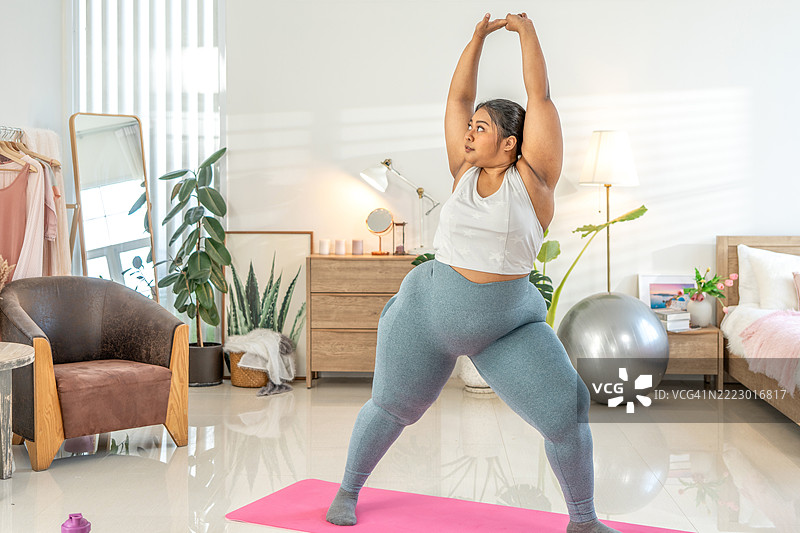 快乐迷人的年轻丰满女性穿着运动服，坐在垫子上，双手放在光脚上，正在做蝴蝶瑜伽动作，拉伸大腿。身体形态与活动。图片素材